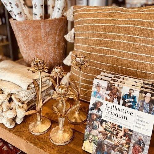 Beige boho tabletop display with gold candle holders, a woven pillow, and a “Collective Wisdom” magazine.