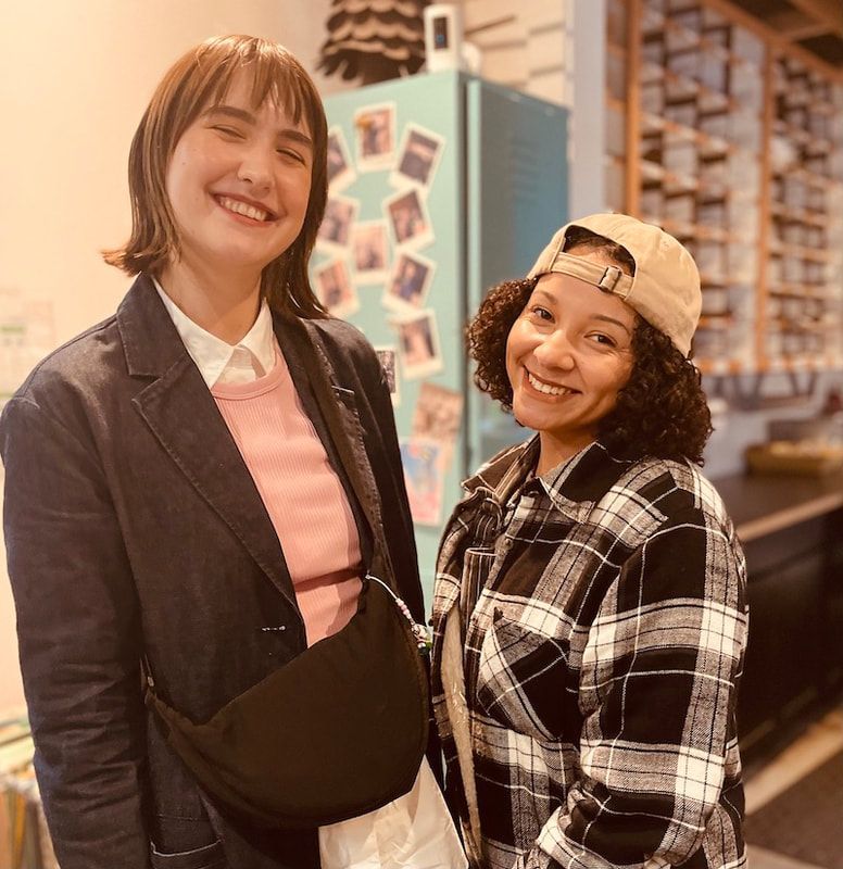 Two smiling people standing indoors, one in a blazer and the other in a plaid shirt and cap.