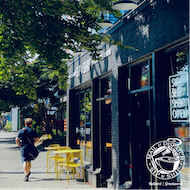 Tree-lined sidewalk with a person walking past a black storefront and yellow outdoor chairs.