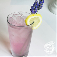 Pink cocktail with ice, lemon slice, and lavender garnish on a white table
