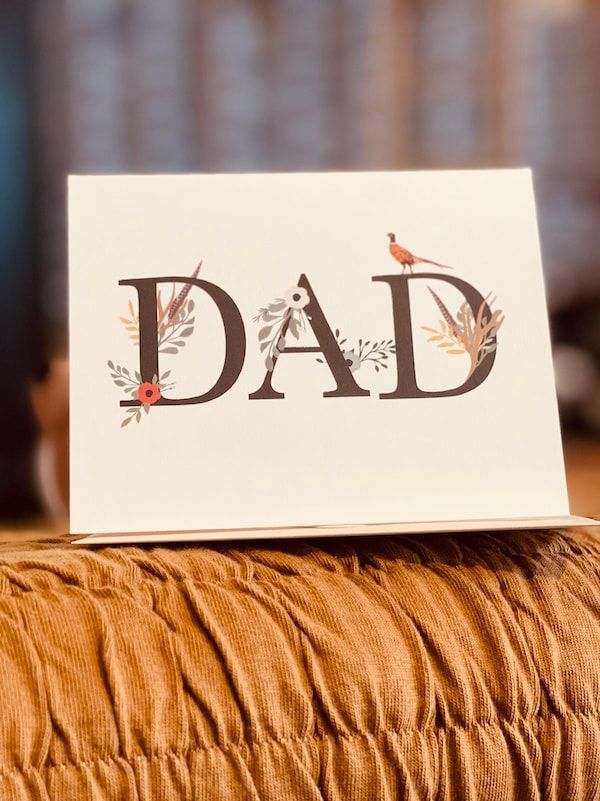A card reading “DAD” with small floral accents, standing on a brown fabric surface.