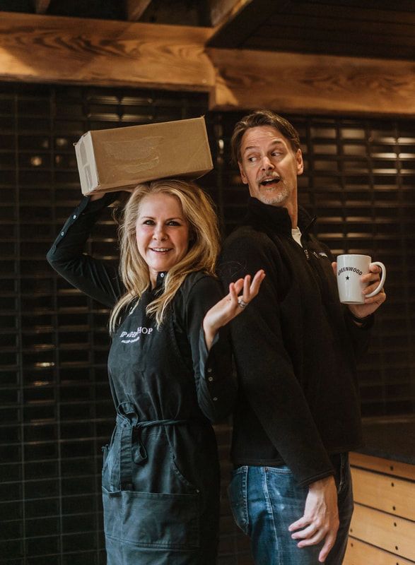 Two smiling people in dark aprons pose indoors, one balancing a box and the other holding a mug.