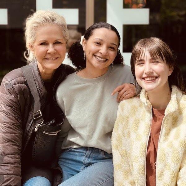 Three smiling friends sitting close together outdoors, wearing casual clothes in front of a building.
