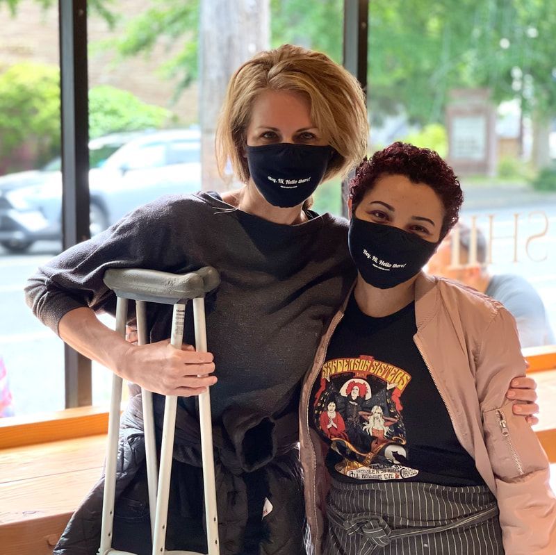 Two people wearing black masks pose indoors, one holding crutches, with large windows behind them.