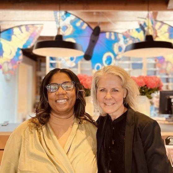 Two smiling people standing together in a brightly lit café, with colorful wall art and hanging lamps behind them