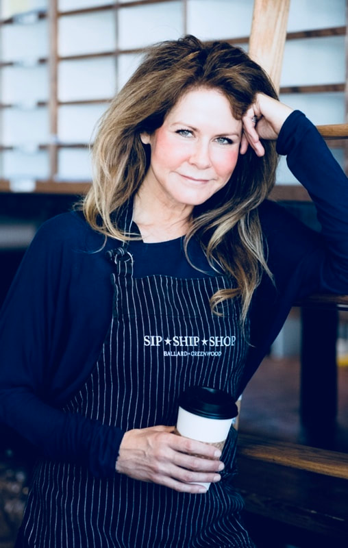 Woman leaning on a counter, holding a white coffee cup, wearing a dark cardigan and striped shirt indoors.