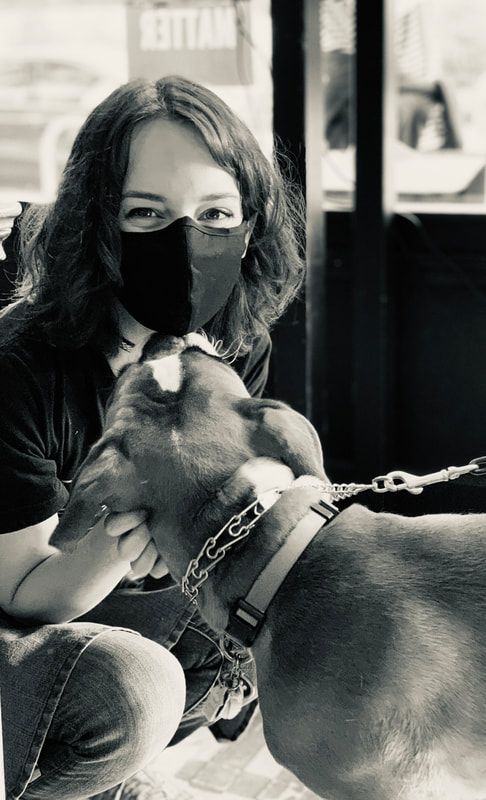 Person wearing a black mask sits with a dog in a harness indoors