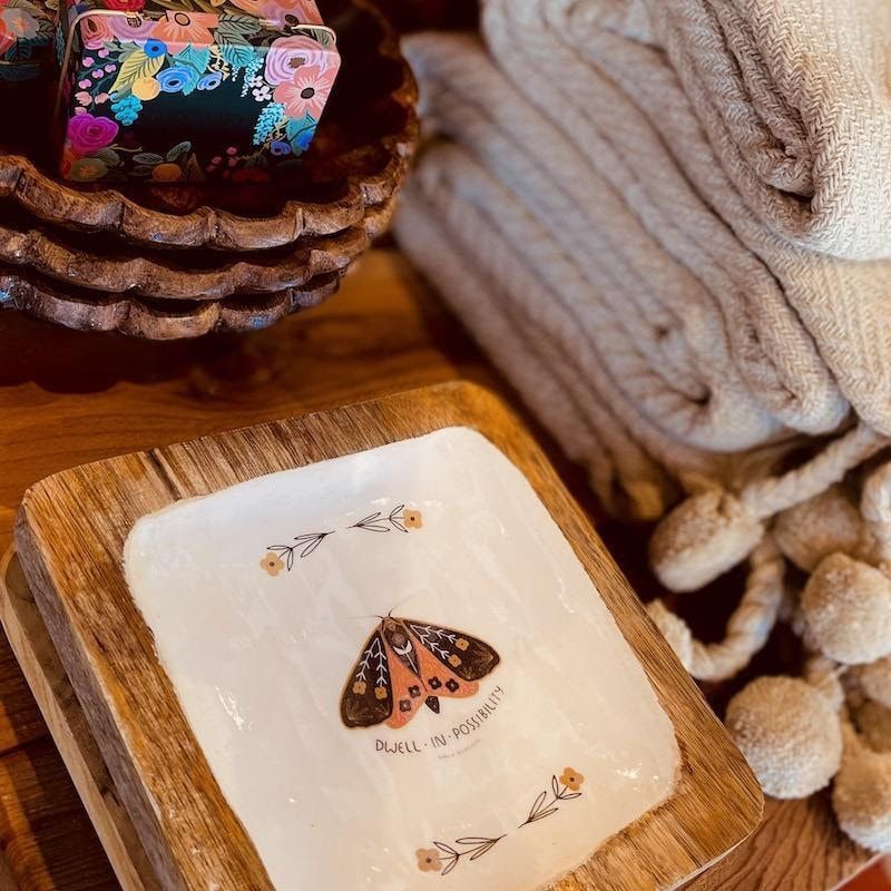 Assorted decorative trinkets: butterfly tile, woven bowls, floral cup, and folded beige fabric on a wooden surface