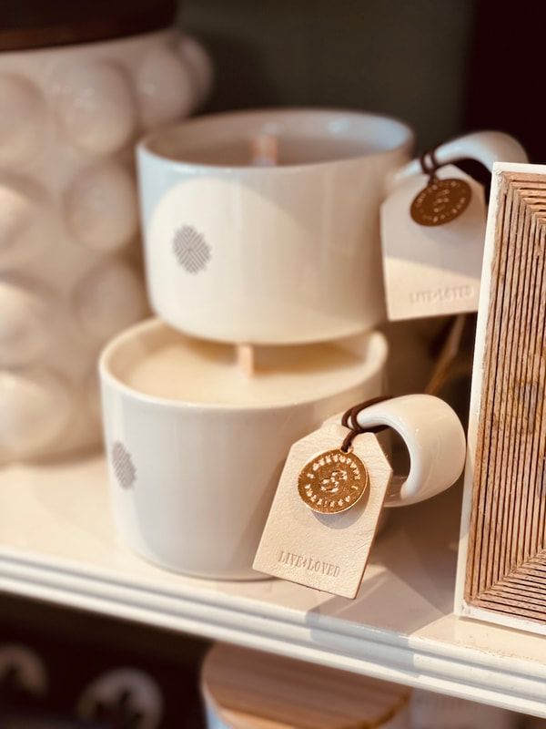 Stacked white mugs with saucers and hanging tags on a shelf, beside a woven container.