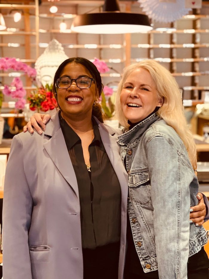 Two smiling people posing together in a flower shop, one in a lavender blazer and one in a denim jacket.