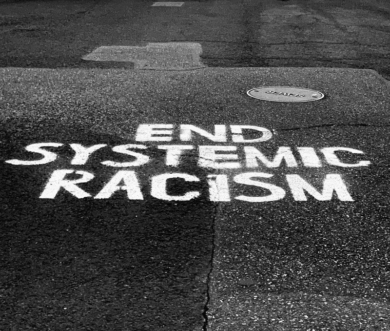 White street graffiti reading “END SYSTEMIC RACISM” on dark pavement.