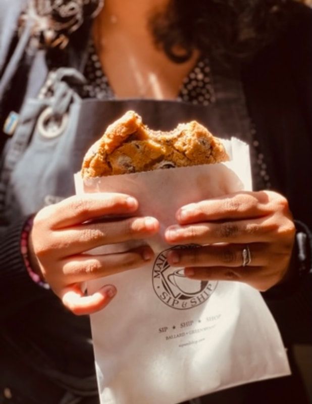 Hands holding a pastry in a white paper bag, with a blurred backpack and warm sunlight in the background