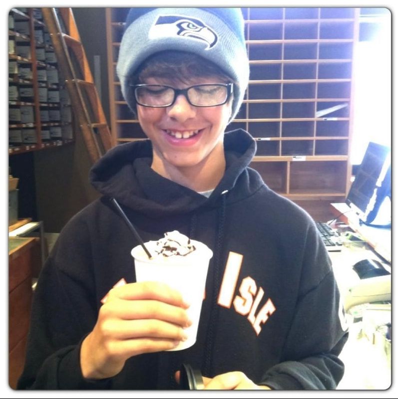Person smiling and holding a white drink in a shop, wearing a gray cap, glasses, and black hoodie.