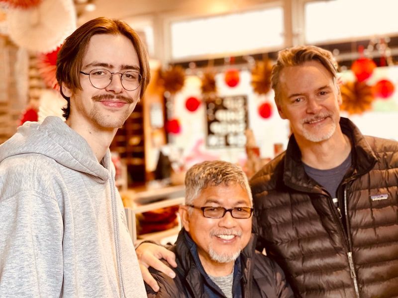 Three people smiling indoors, one with an arm around the others in a warmly lit setting