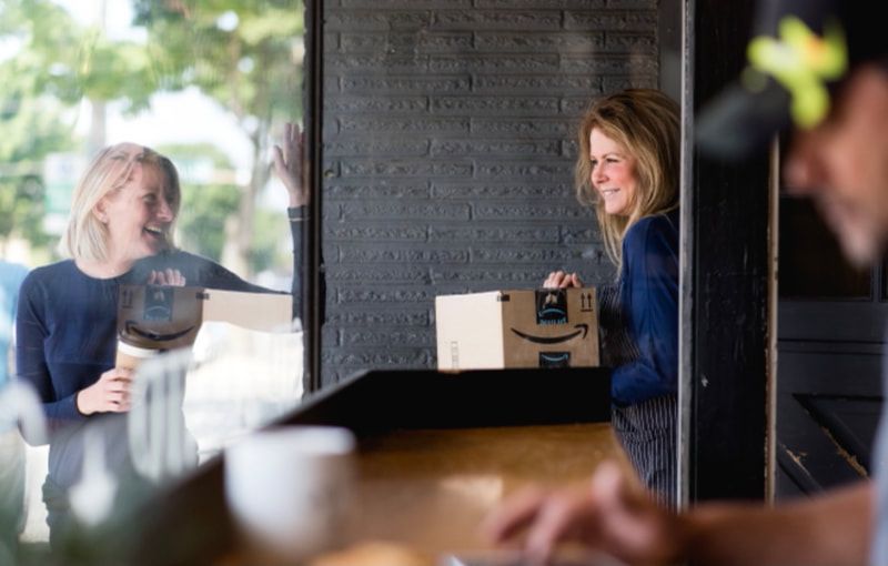 Two people chatting across a counter in a dim café, one holding a drink near a bright window.