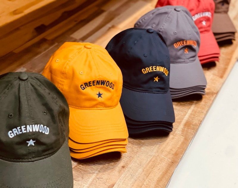 Assorted Greenwood baseball caps in green, yellow, navy, gray, and red displayed on a wooden table.