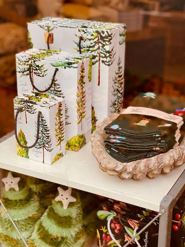 Holiday gift boxes and folded cards on a white display table, with green tree decorations below
