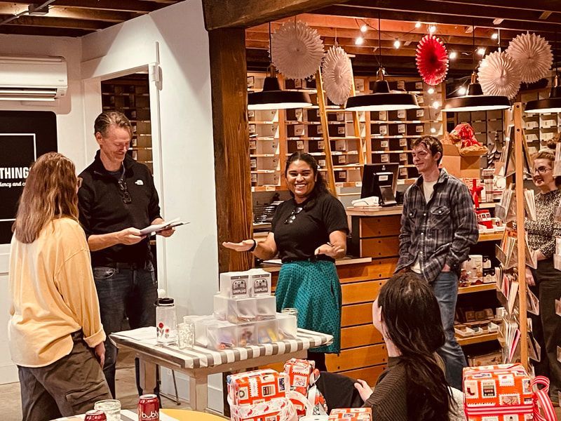 People chatting and browsing in a warmly lit retail shop with shelves and gift displays.