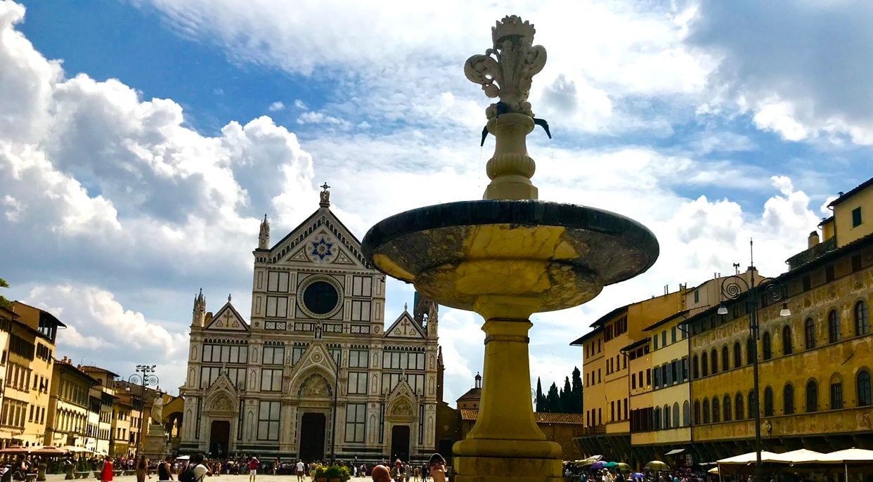 Sunlit cathedral square with a large fountain in front of ornate historic buildings and blue sky with clouds