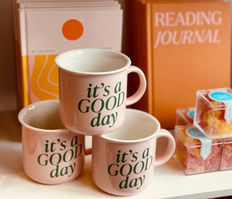 Stacked cream mugs with “it’s a GOOD day” text on a desk beside a reading journal and books.