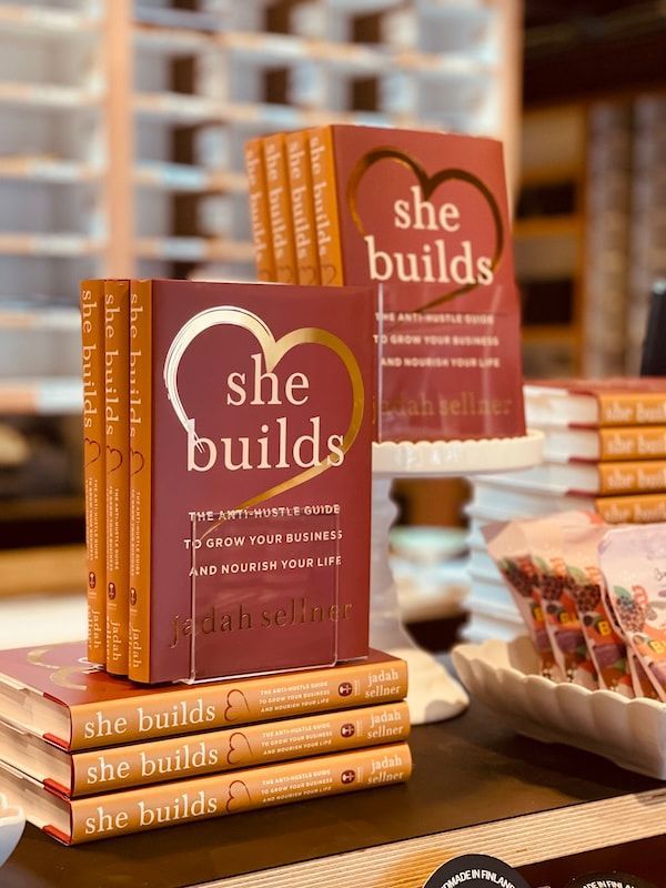 Stacked orange “She Builds” books displayed on a table at a book event booth.