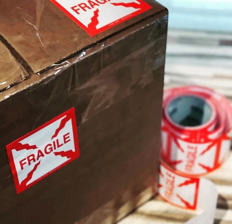 Brown shipping box with red “FRAGILE” stickers beside a roll of fragility tape