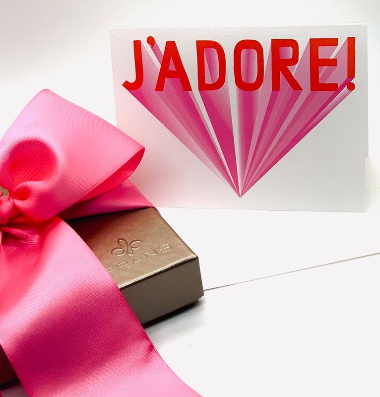 Gift box with pink ribbon beside a card reading “J’ADORE!” in red, with a pink fan design