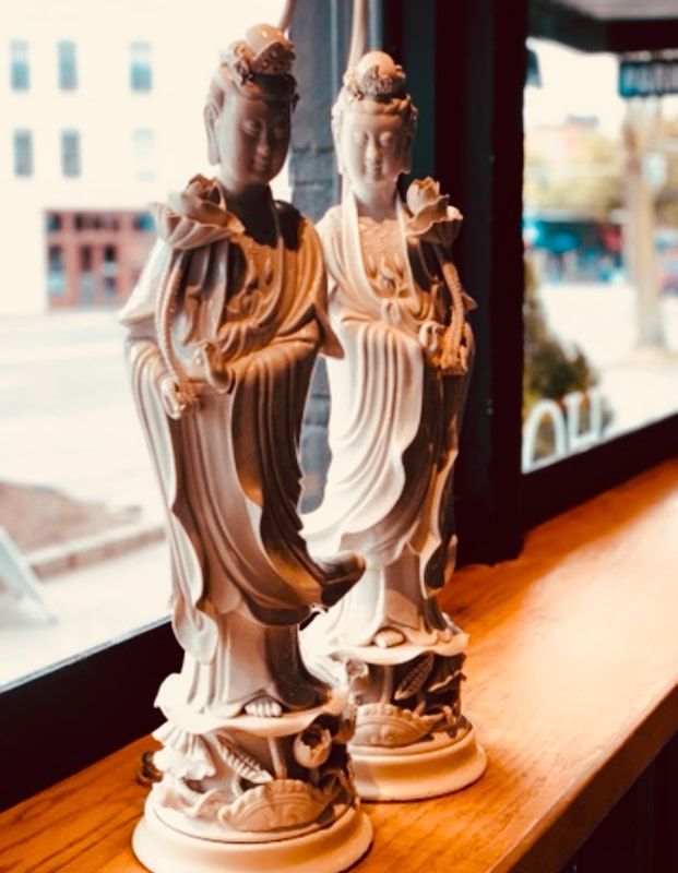 Two ornate white figurines on a wooden windowsill beside a bright window.