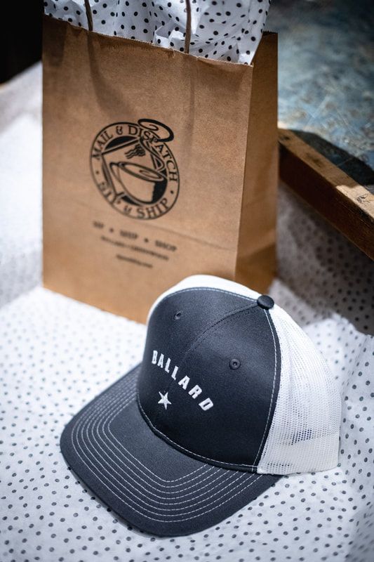 Black and white Vallard baseball cap beside a brown paper shopping bag on a white dotted surface