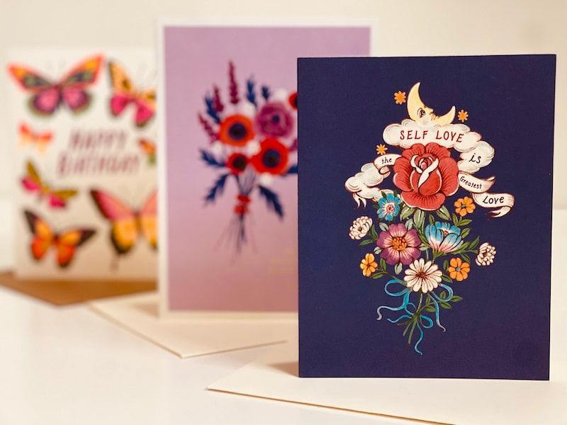 Three floral greeting cards displayed on a shelf, with a navy card in front and butterfly card blurred behind.
