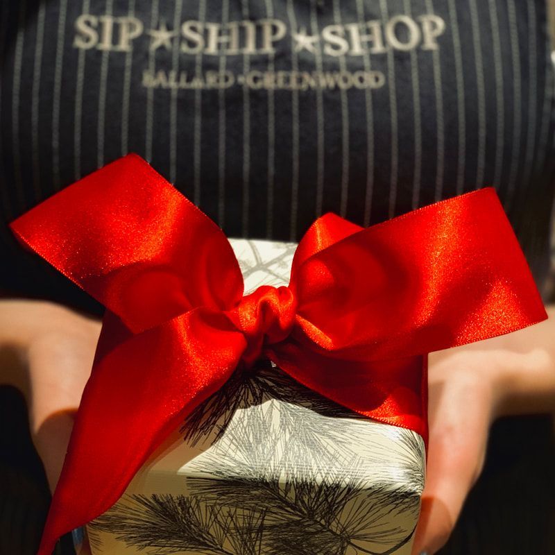 A gift wrapped with a large red ribbon, held in front of a striped apron reading “SIP + SHIP SHOP.”