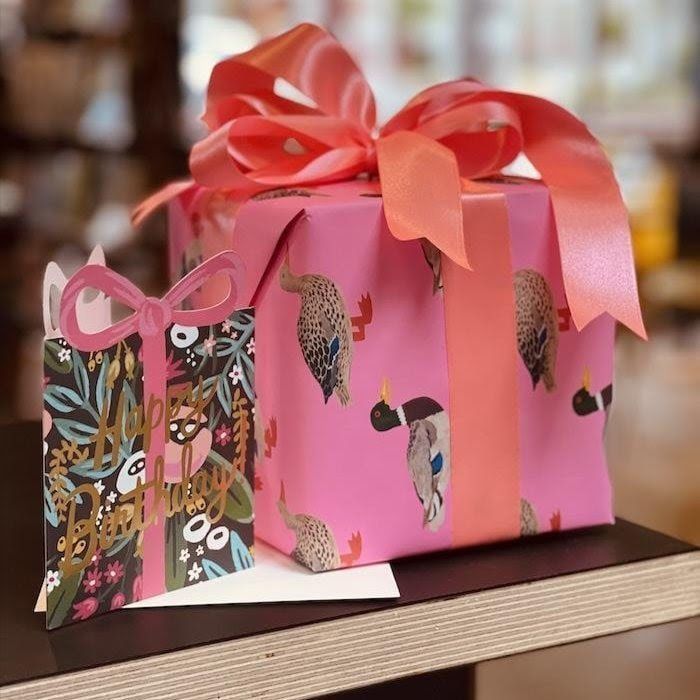 Pink gift box with red ribbon on a wooden surface, beside a colorful patterned gift bag