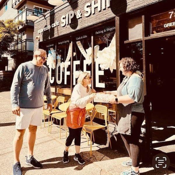 Three people chatting outside a coffee shop, one holding a drink in the sunlight.