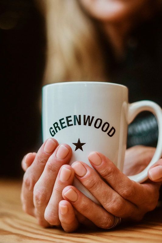 Hands holding a white “GREENWOOD” mug on a wooden table