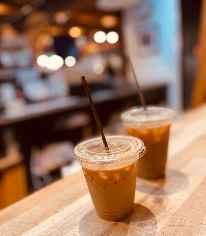 Two iced coffee drinks in clear cups on a wooden table in a warm, blurred cafe setting