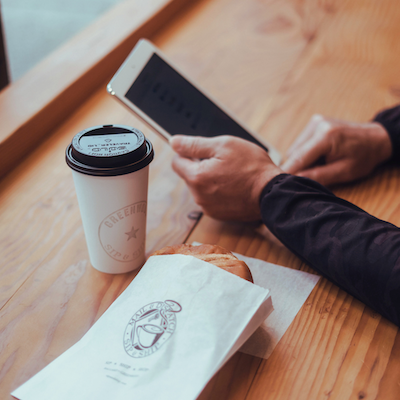 Coffee cup and tablet on a wooden table, with a hand holding the tablet and a paper bag in the foreground