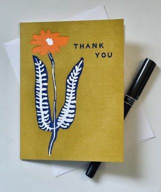 Gold thank-you card with orange flower and blue leaves, next to a black pen on a white surface