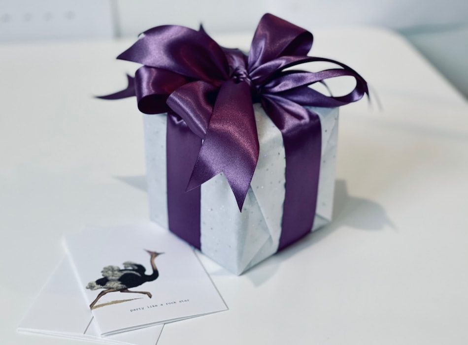 White gift box with a purple ribbon bow on a table, beside a small card with a black illustration