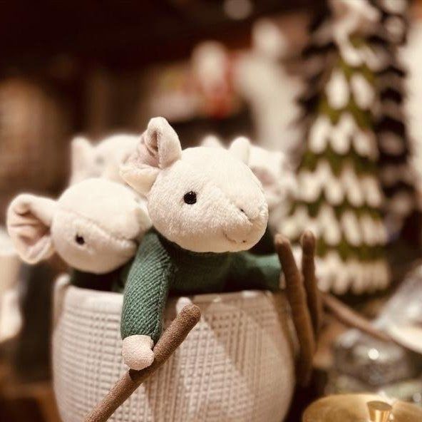 Two plush white mice in a woven basket, one in a green sweater, with a blurred holiday background