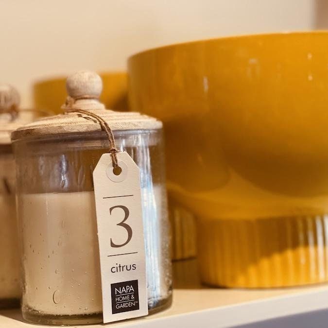 Glass citrus candle jar with tag beside a mustard-yellow container on a shelf
