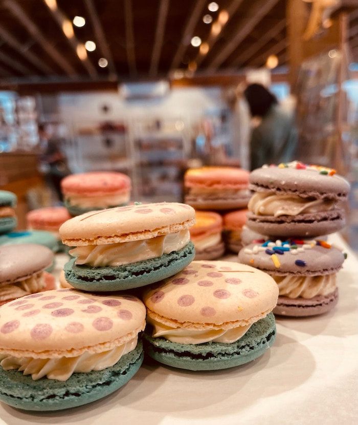 Pastel macarons with polka-dot tops displayed on a bakery counter, with more sweets blurred in the background