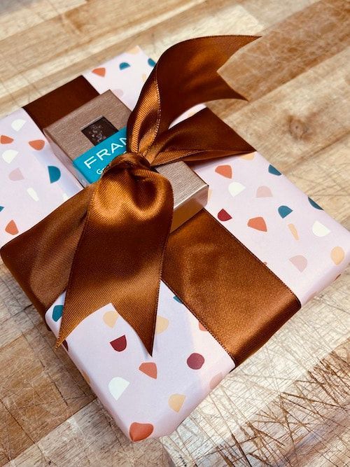Wrapped gift box with white patterned paper and a brown satin ribbon on a wooden surface