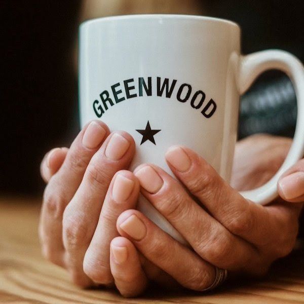 Hands wrapped around a white mug labeled “GREENWOOD” with a star logo