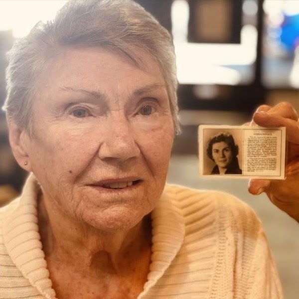 Older woman in a cream sweater holds up a small ID card with a portrait photo and text.