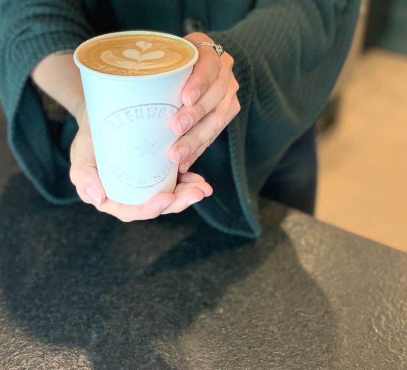 Hands holding a white paper cup of latte with heart latte art on a table.