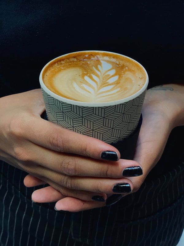 Hands with black nail polish holding a cup of latte with leaf latte art against a dark background
