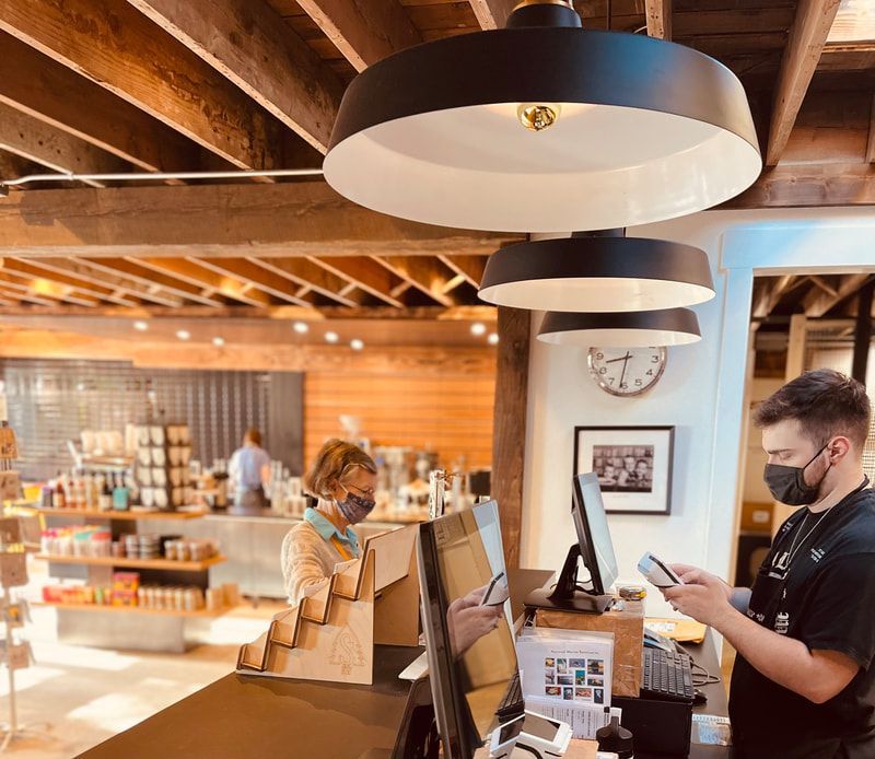 Masked staff at a café counter with laptop and pendant lights, in a warm wood-paneled interior