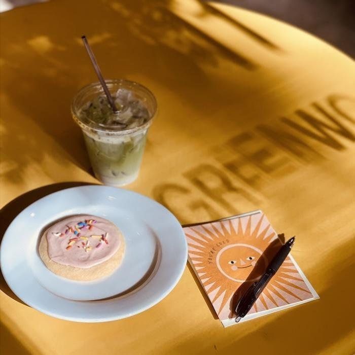 Iced drink, frosted dessert on a plate, and napkin on a yellow table with sunlight shadow.