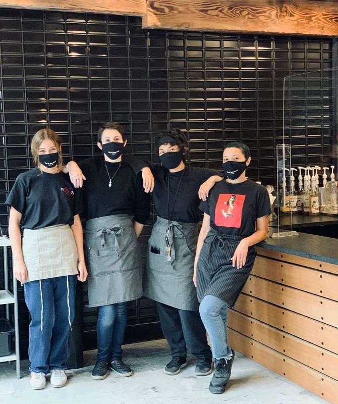 Four masked café staff in aprons stand behind the counter, posing in front of dark shelves.