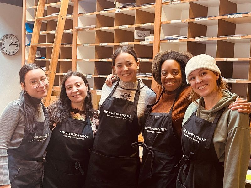 Five smiling people in aprons stand together in front of wooden shelves in a workshop.